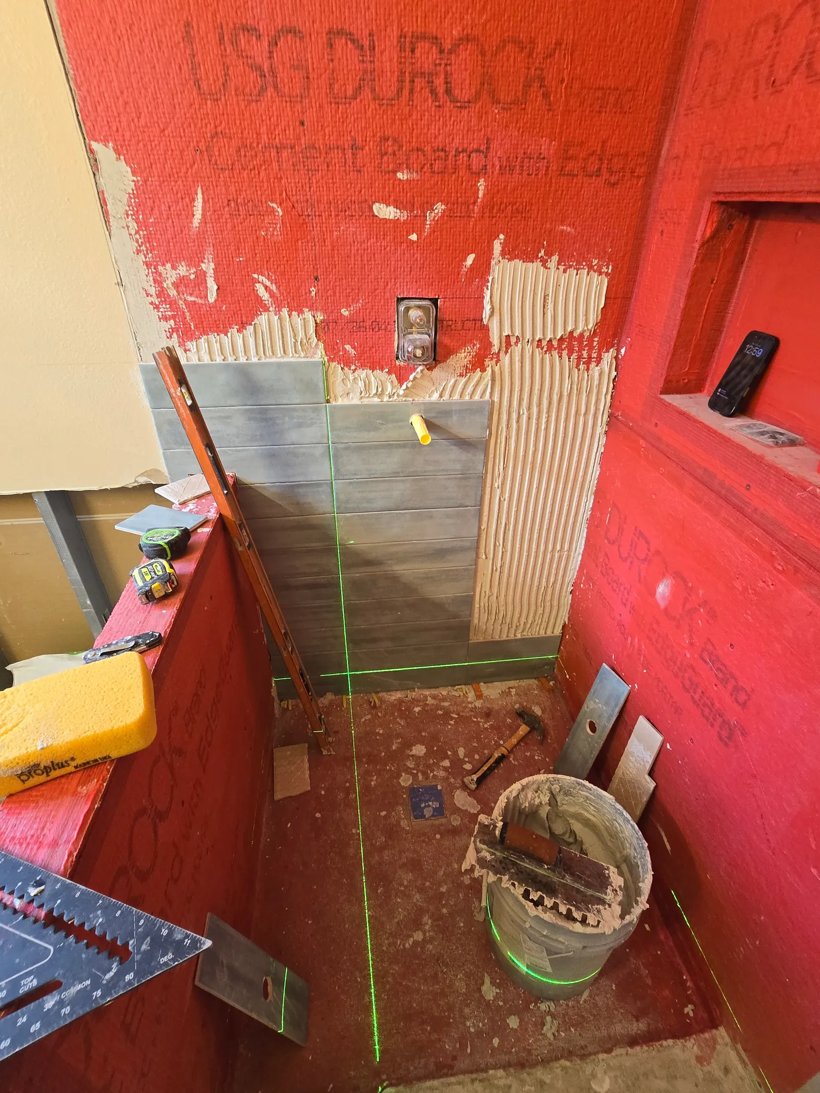 Shower walls in red waterproofing membrane with the first rows of grey tile set with a laser level