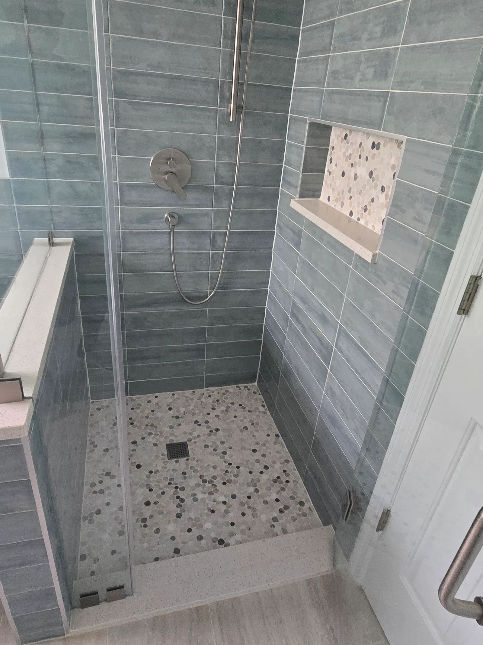 Finished walk-in shower with rain head, hand shower, mosaic niche and pebble floor behind frameless glass